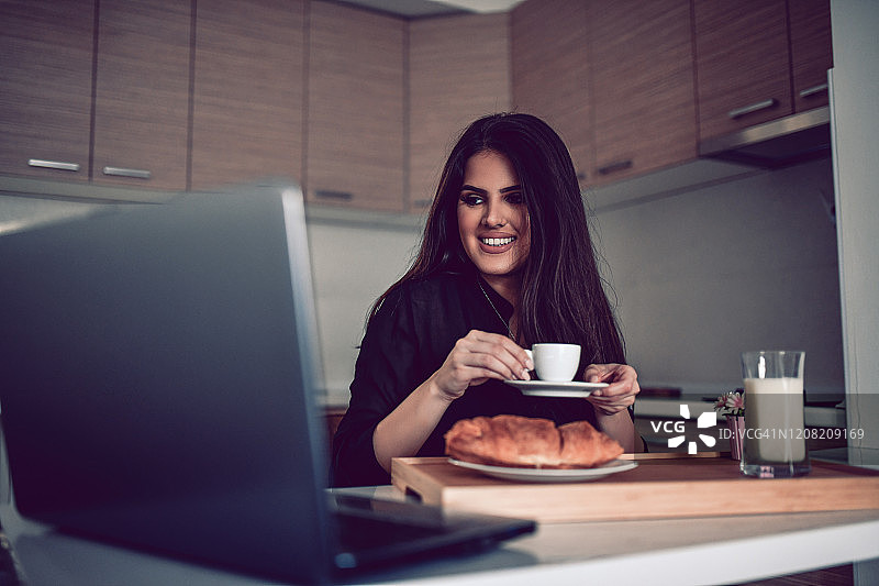 可爱的女人一边喝早咖啡和早餐，一边在笔记本电脑上看视频图片素材