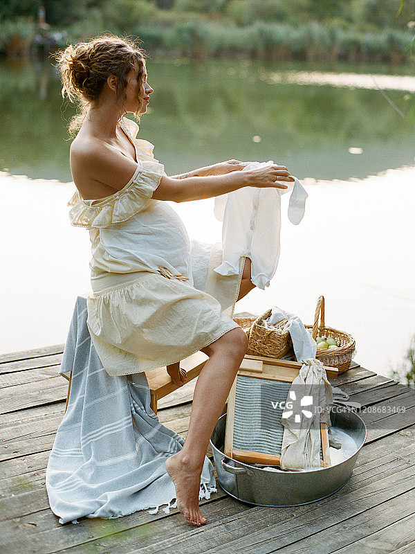 穿着复古连衣裙的年轻孕妇在河边金属盆里清洗亚麻布，孕妇复古海报风照片，生态洗衣概念图片素材