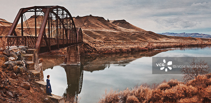 在老铁路桥底的墙上，一位扎着红色辫子，穿着蓝色复古长袖连衣裙的年轻女子背影，俯瞰河流图片素材