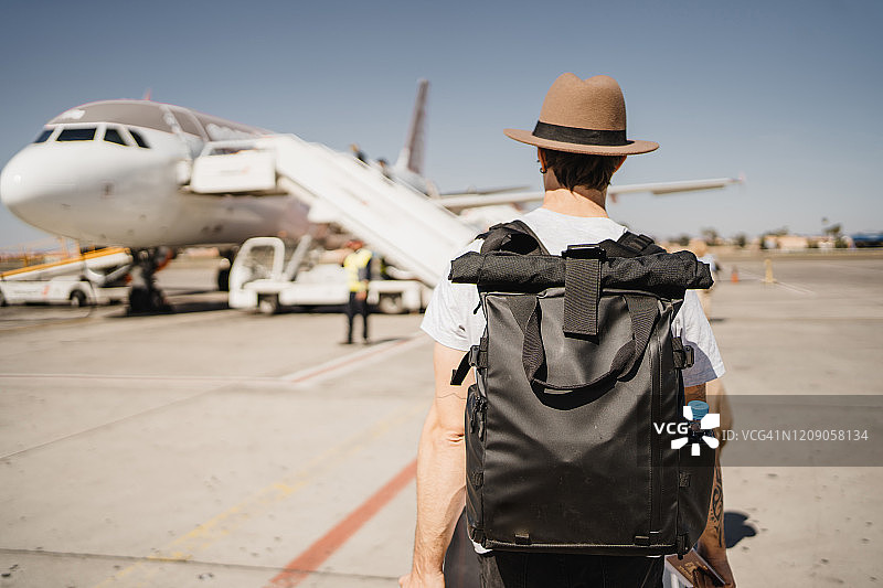 年轻的白人男子戴着帽子，拖着手提箱走在机场跑道上，走向他的飞机图片素材