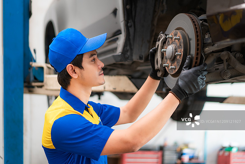 身穿蓝色制服的亚洲汽车修理工正在汽车服务中心检查和修理车辆的盘式制动器。维修、汽车服务和维护概念。图片素材