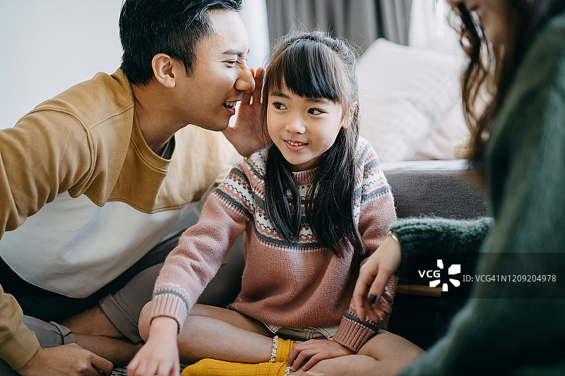 年轻父母与可爱女儿在家中温馨互动，父亲在女儿耳边轻语图片素材