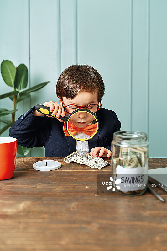 2-3岁儿童穿着西装像个商人一样在他的办公桌上工作，他用放大镜看钱图片素材
