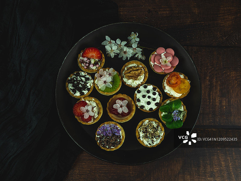 木桌上摆放的圆形黑色托盘，含甜味和咸味小吃及食用鲜花图片素材