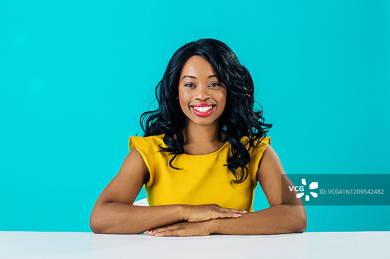 一位快乐的年轻微笑女性坐在办公桌后，双臂交叉，在蓝色背景下隔离图片素材