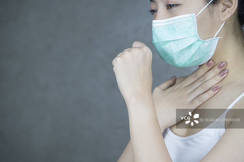 一位亚洲女性感冒咳嗽，戴着防护或卫生口罩的女孩站在浅灰色墙壁背景前，预防病毒。 疾病、冠状病毒概念图片素材