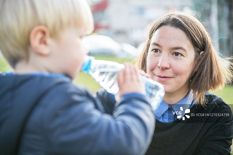 在欧洲游乐场喝水的金发幼儿，母亲在一旁看护图片素材