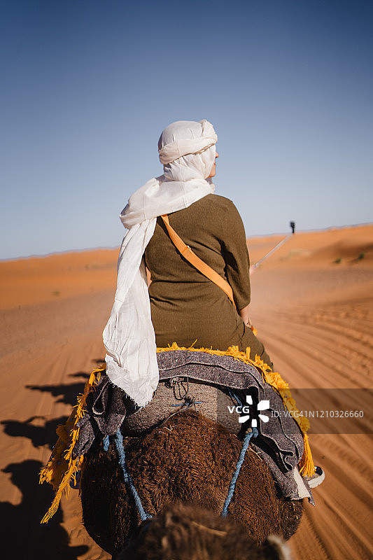 一名戴着白色面纱的女子骑着骆驼穿过撒哈拉沙漠的背影图片素材