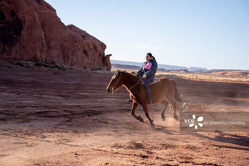 年轻的纳瓦霍族少女骑马飞奔，小狗在后面追赶图片素材