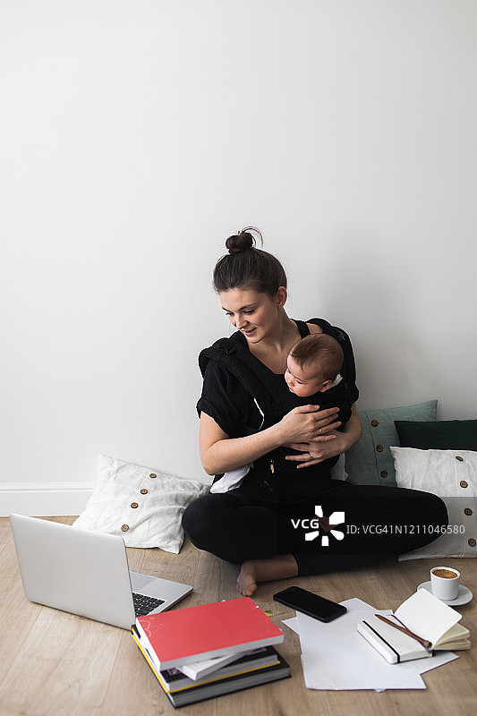 年轻漂亮的母亲抱着婴儿在电脑上工作，在产假期间忙于多任务处理。在家工作。图片素材