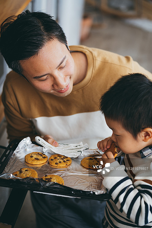 父亲和儿子在厨房一起烤饼干，儿子迫不及待地想品尝刚出炉的饼干图片素材