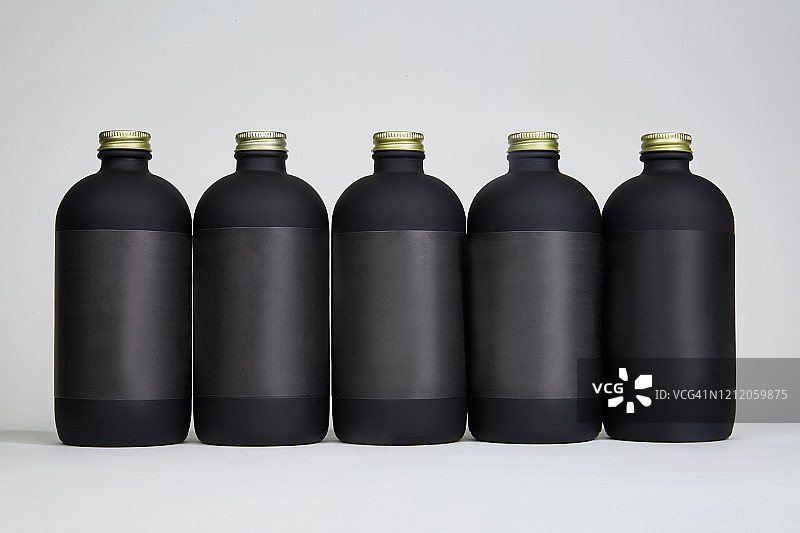五个带黑色标签和金色瓶盖的16盎司哑光黑瓶，灰色背景图片素材