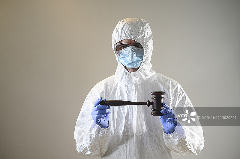 戴着口罩、手套和防护服（生化防护服）的男人双手拿着法官的法槌。平衡、正义和冠状病毒概念。图片素材