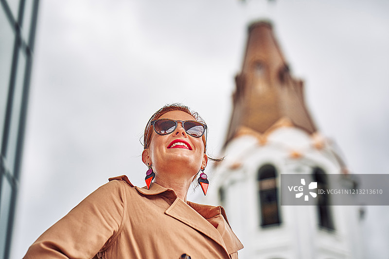 一位身穿红色衬衫、棕色斗篷、戴着眼镜的中年红发女子在城市里散步的肖像。阴沉的夏日。特写。图片素材