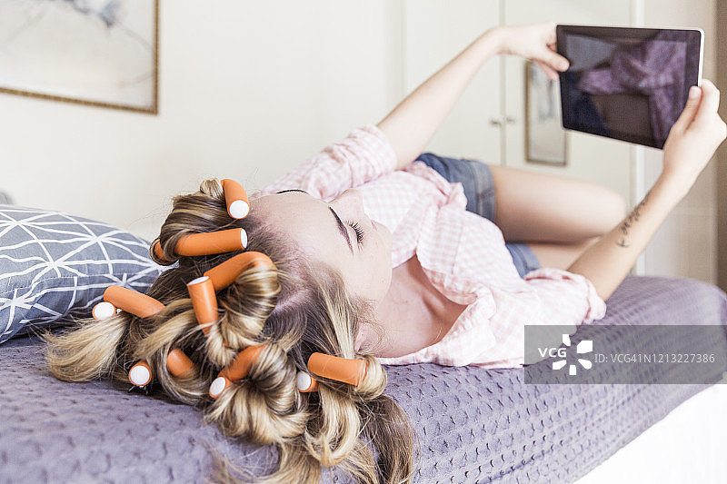 躺在床上用平板电脑自拍的年轻女子，头发上卷着发卷图片素材