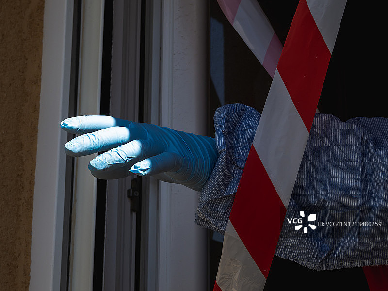 在2019年新冠病毒危机期间，男人戴着防护手套从带有警戒线的窗户伸出手图片素材