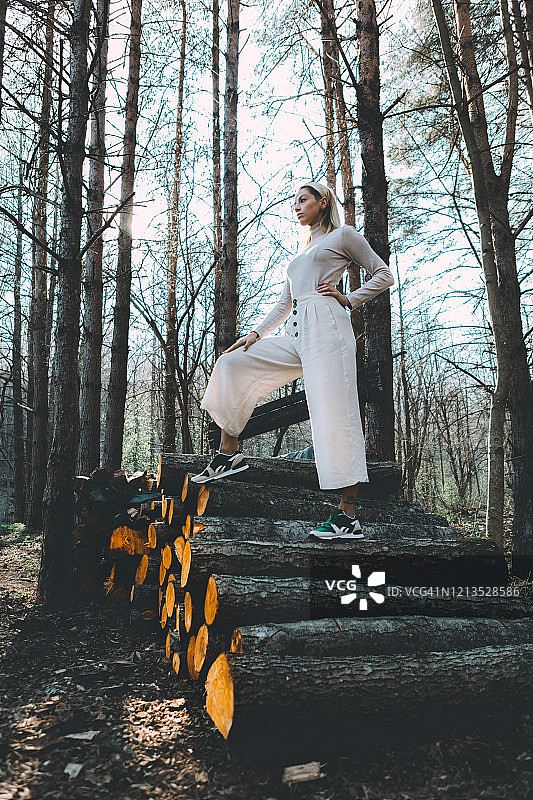 站在木墙前的金发长发女子，身穿白色亚麻毛衣和白色阔腿裤，头戴发带，阳光穿过树林图片素材