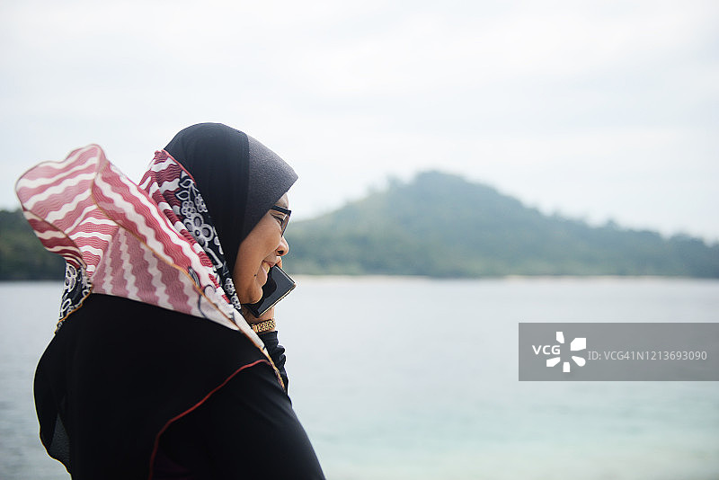 在海滩上使用智能手机的戴头巾的亚洲穆斯林女人图片素材