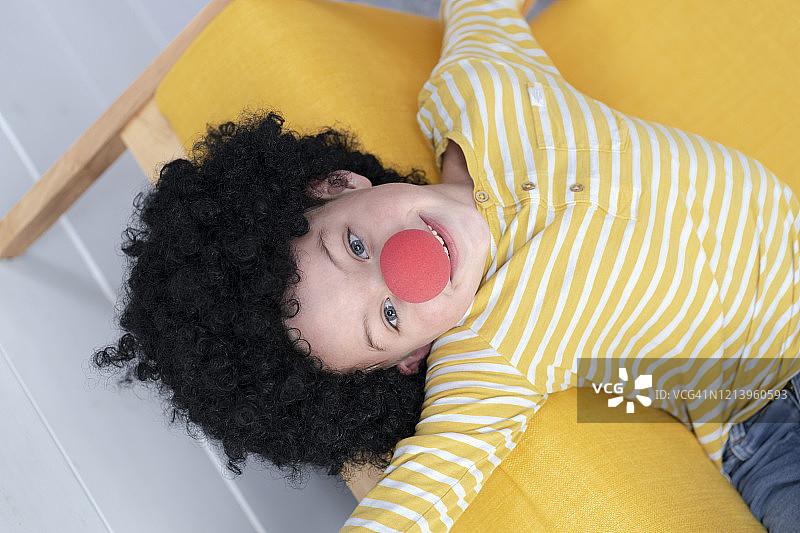 戴着红色小丑鼻子，坐在黄色沙发上的黑发男孩图片素材