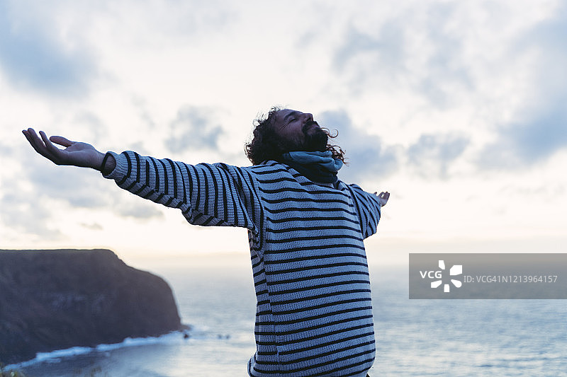 日落时分，男子在葡萄牙亚速尔群岛圣米格尔岛海岸张开双臂站立图片素材