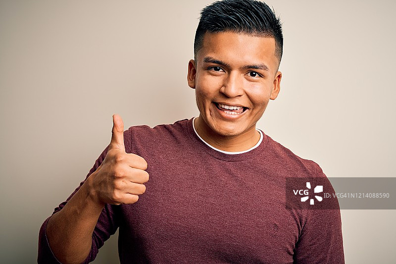 在白色背景下手持大拇指的年轻英俊拉丁男子，面带微笑图片素材