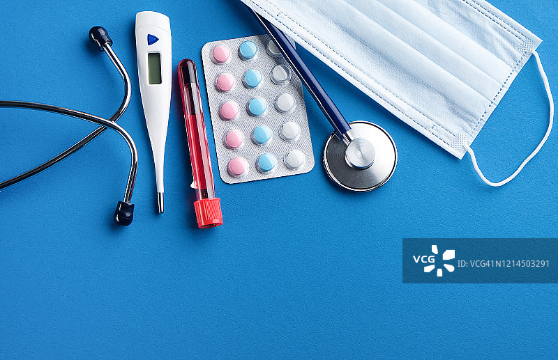 蓝色背景上的医用一次性防护口罩、温度计、药片和药物、听诊器和试管中的血液样本图片素材