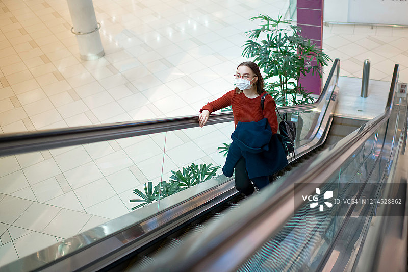 在购物中心自动扶梯上戴着医用口罩以保护自己免受病毒或污染的年轻女孩图片素材