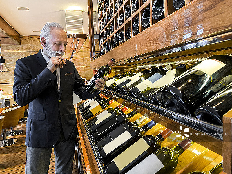 年长的侍酒师在豪华酒窖中挑选葡萄酒，并抽着雪茄图片素材