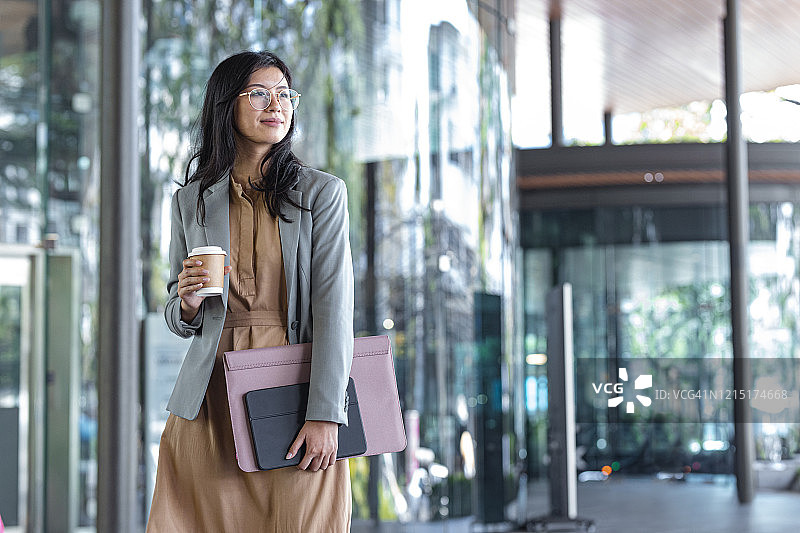 快乐的亚洲女商人手拿咖啡杯和文件站在玻璃大楼旁图片素材