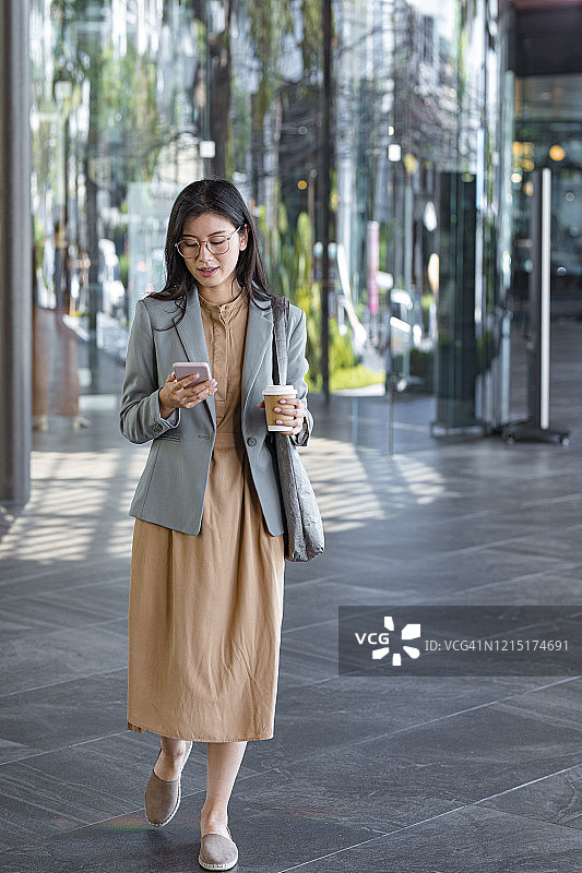 优雅亚洲女性在街上行走并用智能手机发短信全身照图片素材