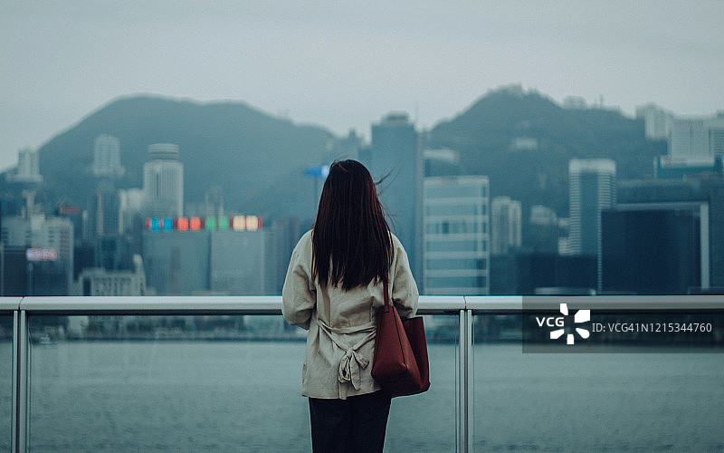 阴天时，女子站在维多利亚港海滨长廊，俯瞰香港城市景观的背影图片素材