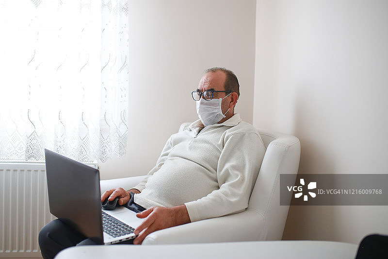 戴口罩的老年男人在客厅用笔记本电脑看电影，居家隔离，新冠疫情期间的生活图片素材