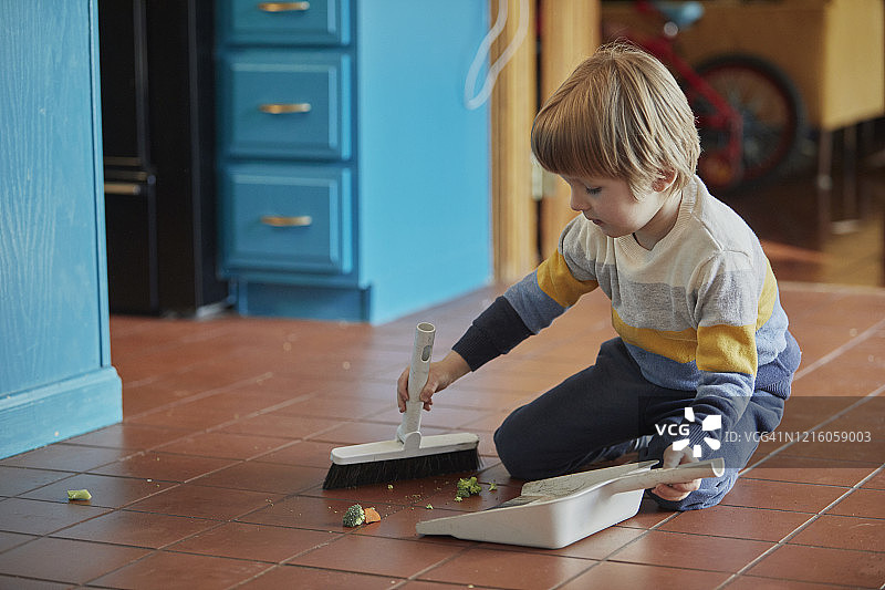 小男孩跪在瓷砖地板上用簸箕和刷子打扫卫生图片素材