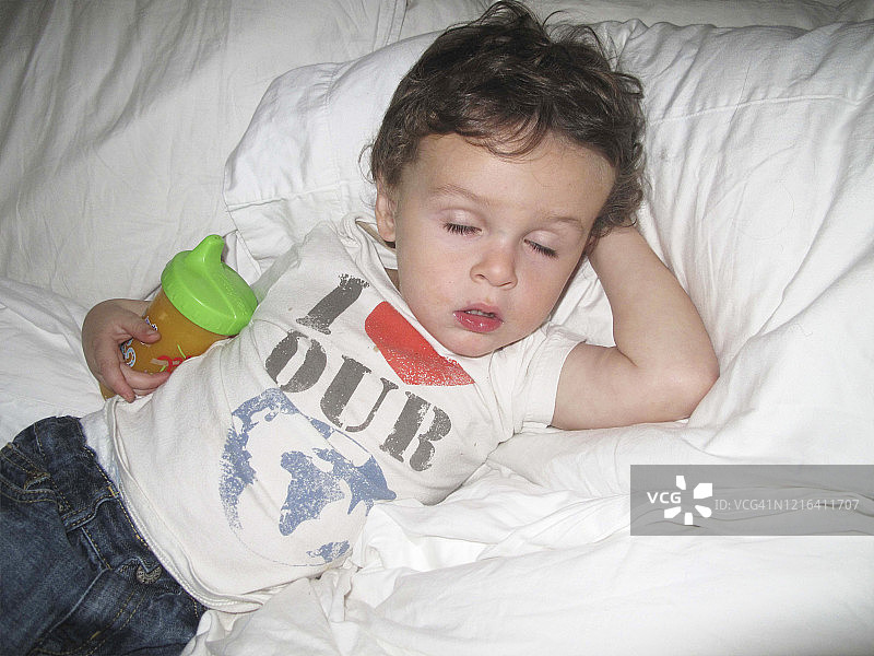 幼儿男孩穿着牛仔裤和T恤，在白色床上睡觉，手里拿着一杯橙汁图片素材