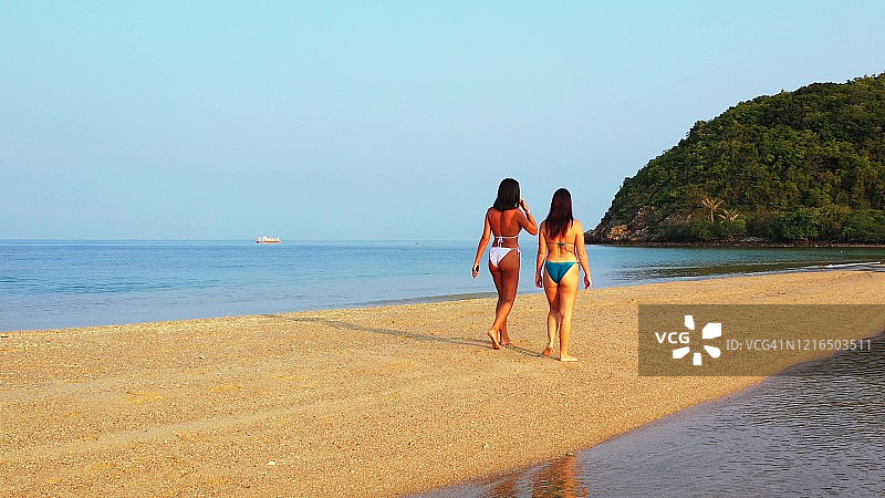 两个年轻女人在泰国沙滩上，平静的浅水泻湖在美丽的晨光下拍打着沙滩图片素材