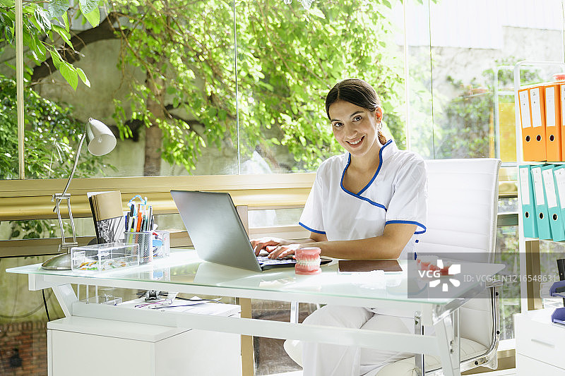 20多岁的牙医在阳光明媚的办公室里用笔记本电脑打字图片素材