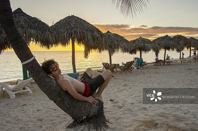 在古巴安孔海滩日落美景中，戴着太阳镜的青少年男孩在树干上放松图片素材