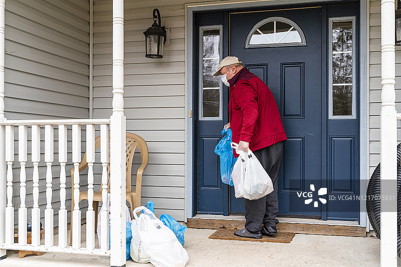 疫情期间，戴口罩和手套的老年男人将食品从门廊的垃圾箱中取出放入家中。图片素材