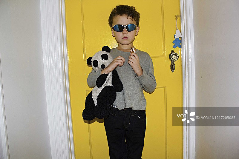 棕色卷发棕色眼睛的小男孩抱着毛绒熊猫玩具，戴着游泳镜站在黄色门框前图片素材