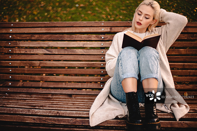 年轻女人在秋天的公园里坐在长椅上读书图片素材