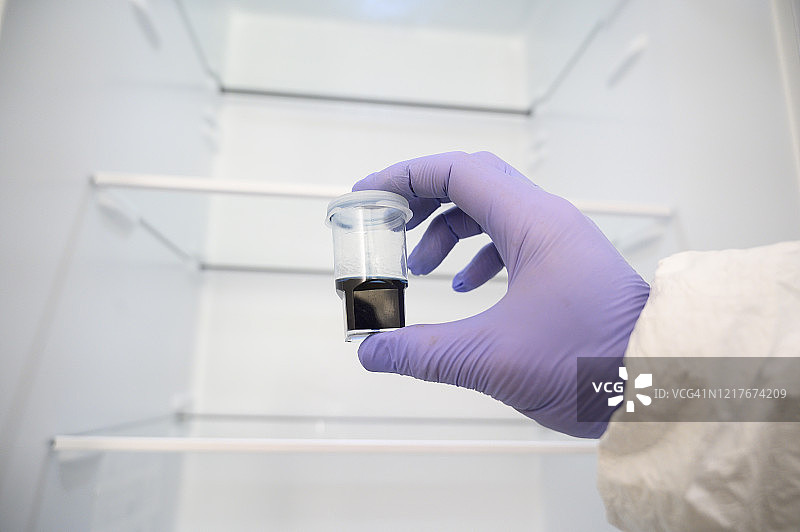 科学家戴着防护手套在实验室里拿着一个医学样本，前面是一个空冰箱。他穿着防护服（危险品防护服）图片素材