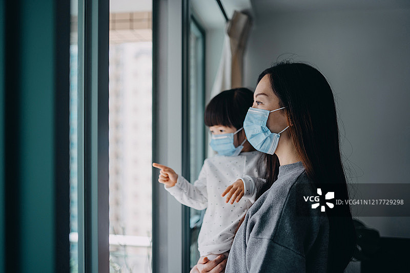 年轻母亲和女儿戴着防护口罩看着窗外，因新冠疫情在家自我隔离图片素材