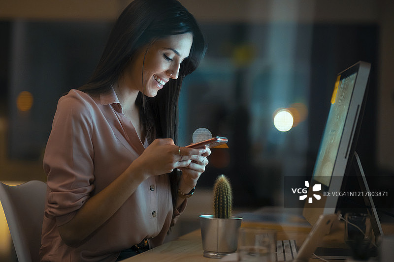 微笑的年轻女人在办公室的桌子旁使用智能手机图片素材