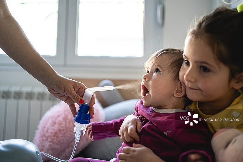 母亲和姐姐帮助过敏的婴儿在家用雾化器图片素材