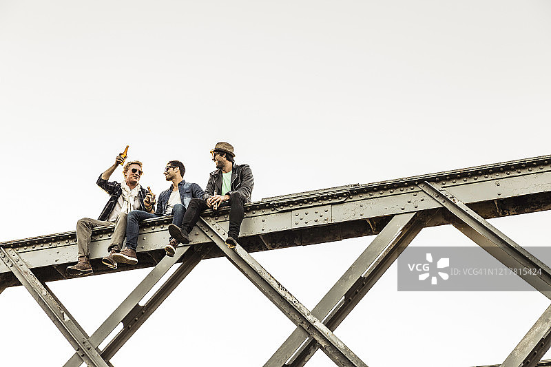 三个男人坐在旧铁路桥的金属杆上喝啤酒图片素材
