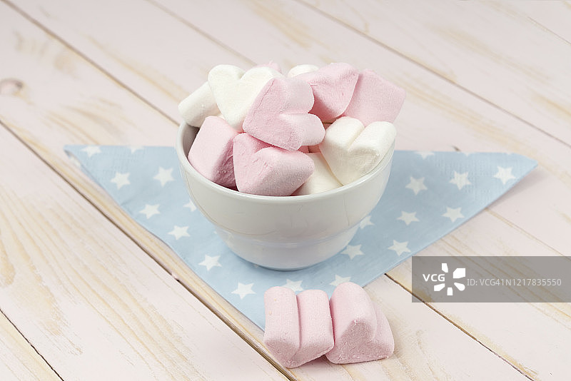 瓷碗里的白色和粉色心形棉花糖，蓝色餐巾置于粉色木纹背景上图片素材