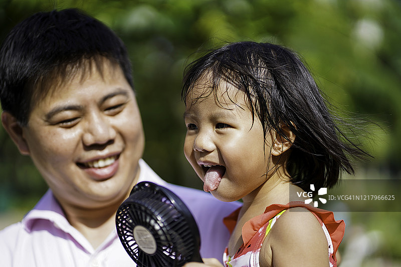 年轻的亚洲父亲在炎热的天气里帮助女儿用便携式风扇扇风图片素材