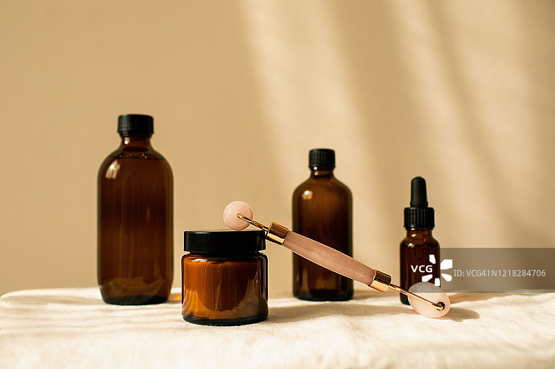 琥珀色玻璃瓶，用于化妆品、天然护肤品、精油芳香疗法或其他液体，配有玫瑰石英面部滚轮，米色背景。草药替代医学。留白图片素材