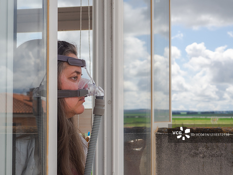 疫情期间，戴呼吸器、站在窗边的中年妇女肖像图片素材
