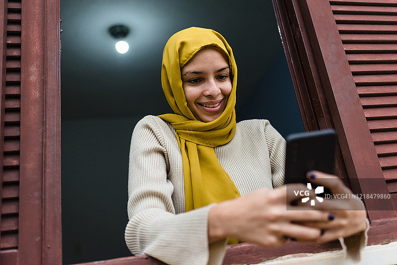 在窗边使用智能手机的穆斯林年轻女人图片素材